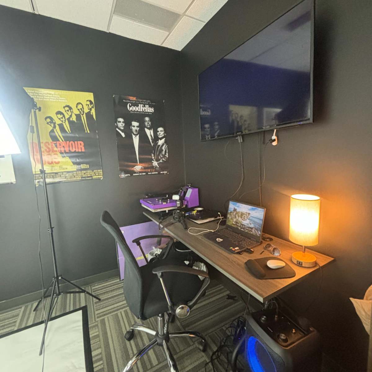 A workspace with a desk, computer setup, lamp, and posters on the wall in a dark-colored room.