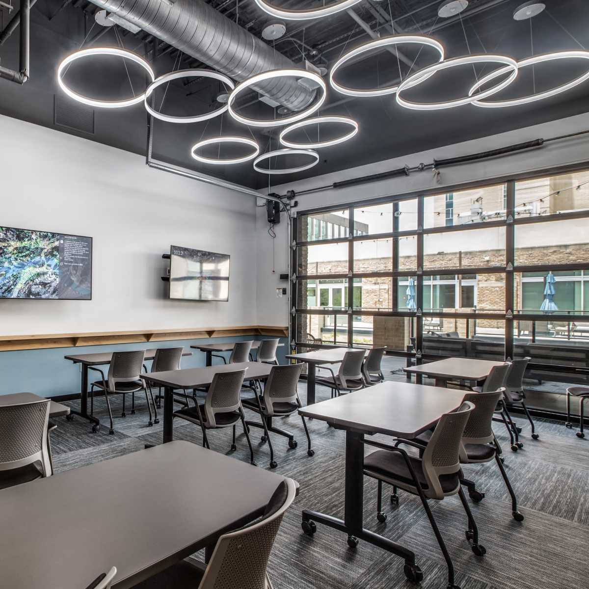 A modern classroom with several movable desks arranged for collaboration, featuring two wall-mounted screens and circular light fixtures overhead.