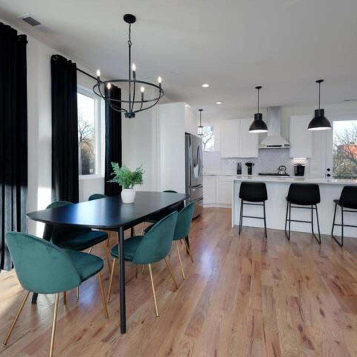 A modern kitchen and dining area featuring a rectangular black table with green chairs, a kitchen island with bar stools, and large windows allowing natural light.