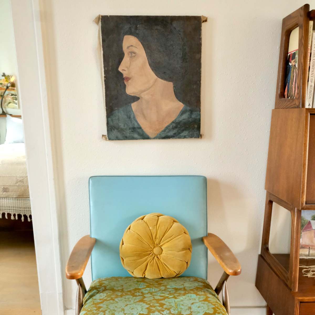 A vintage chair with patterned upholstery and a round cushion sits in front of a portrait of a woman mounted on the wall.
