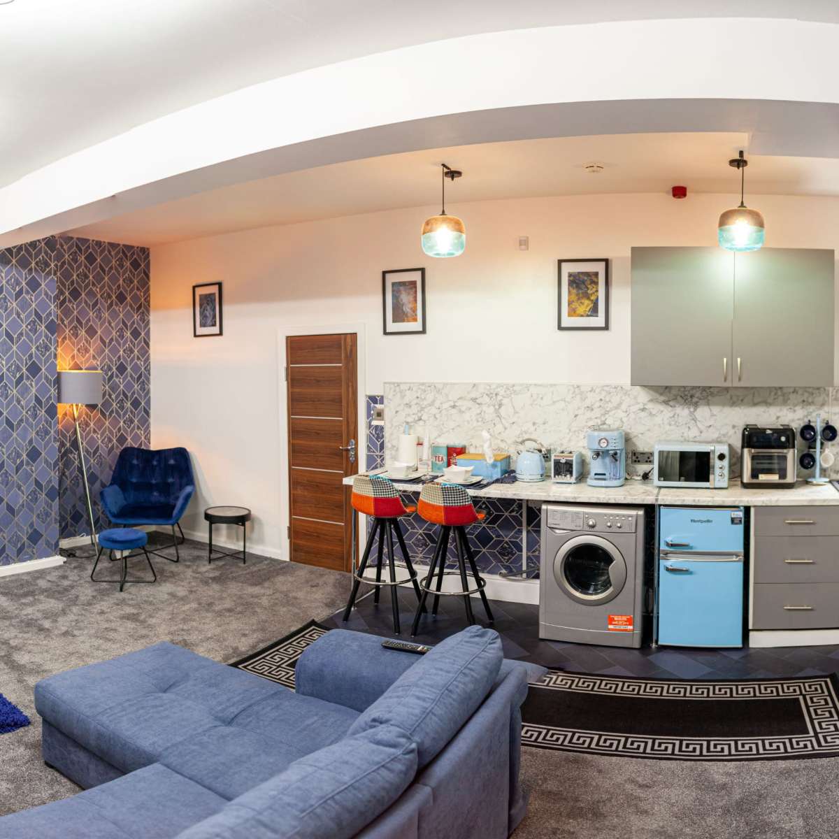 A modern studio apartment with a living area, kitchen, and workspace, featuring blue accents and contemporary furnishings.