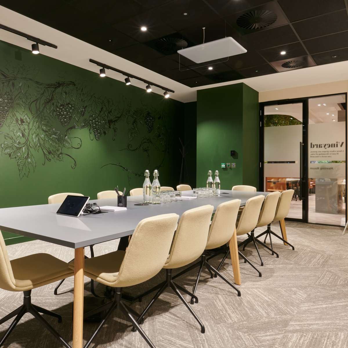 A modern conference room features a long table with beige chairs, green walls adorned with a vine mural, and large windows allowing natural light.