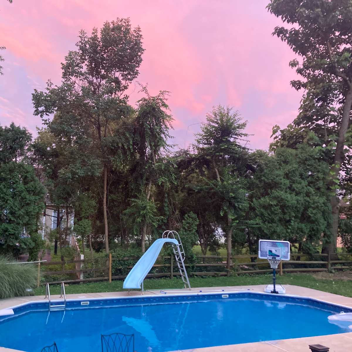 The scene features a backyard pool surrounded by trees and a slide, with a pinkish sky at sunset.
