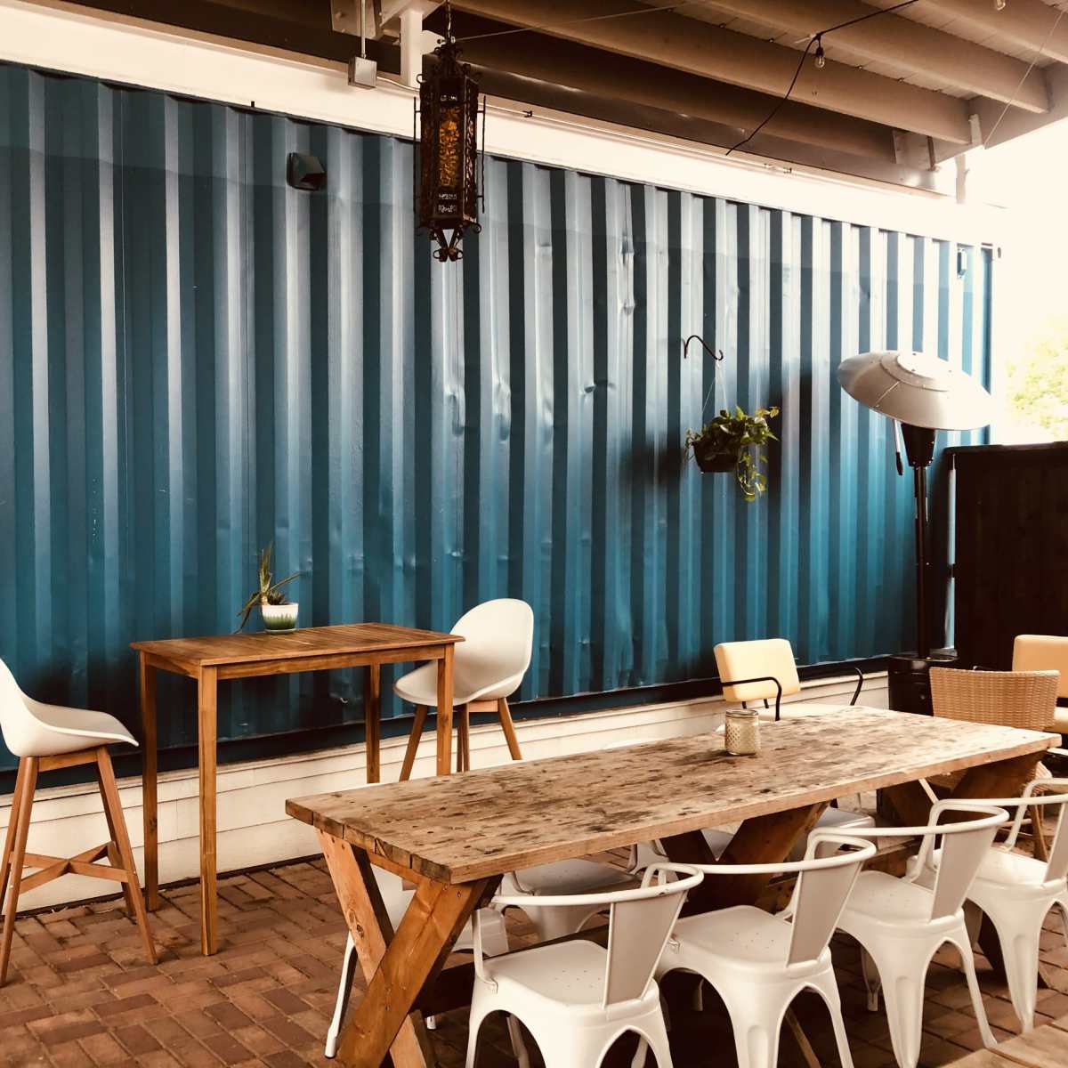A patio area featuring a wooden table and chairs in front of a blue corrugated wall, with some potted plants and an umbrella.