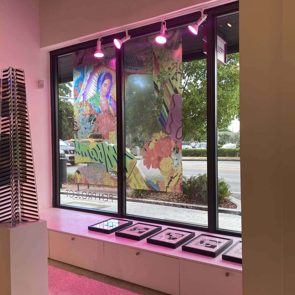 A window display with colorful artwork featuring floral designs and a figure, along with framed pieces below illuminated by pink lights.