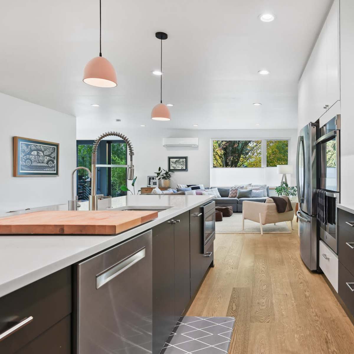 A modern kitchen features dark cabinets, light countertops, and stainless steel appliances, leading into a bright living area with a sofa and large windows.