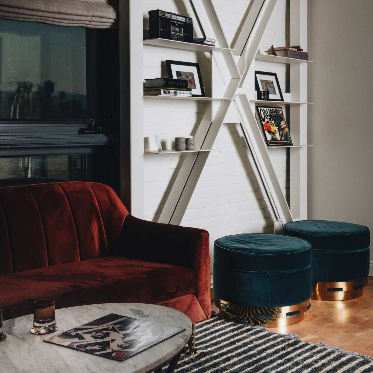 A red velvet sofa and two teal ottomans are positioned near a marble coffee table, against a wall featuring a modern shelving unit and framed photographs.