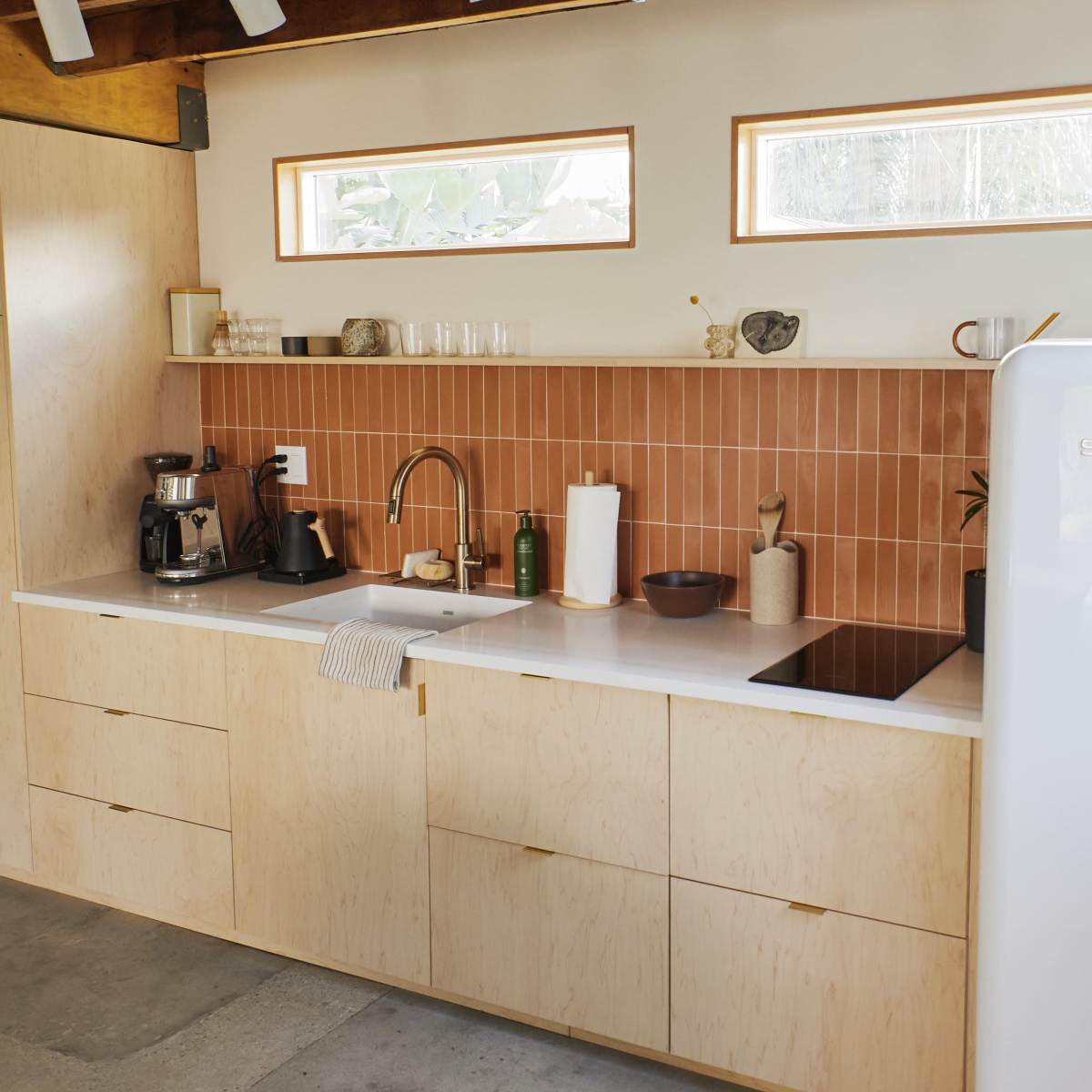 A modern kitchen features a light wooden cabinetry, a white SMEG refrigerator, and a pink tile backsplash with various kitchen essentials on the counter.