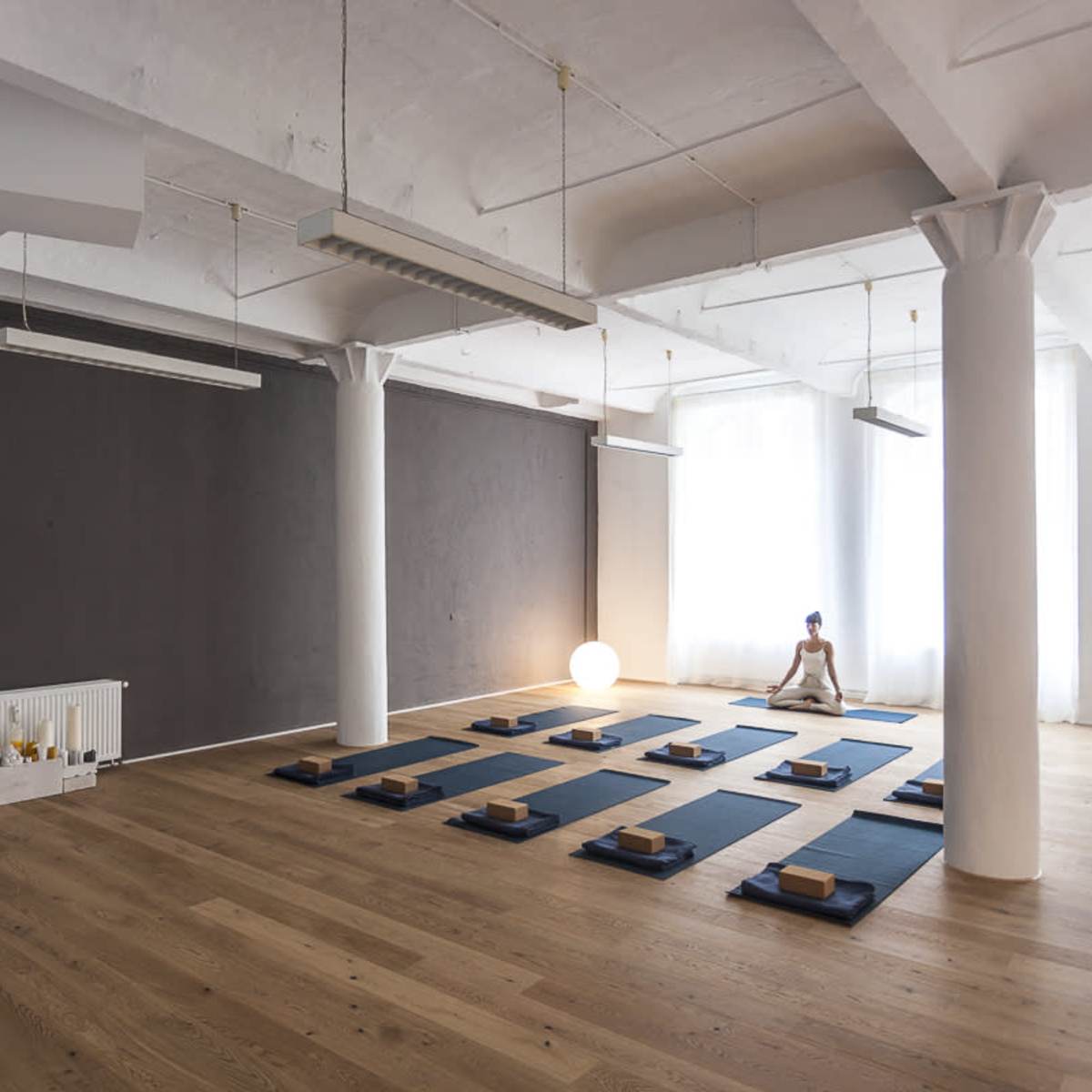 A spacious yoga studio with wooden flooring, white pillars, and exercise mats arranged neatly on the floor.