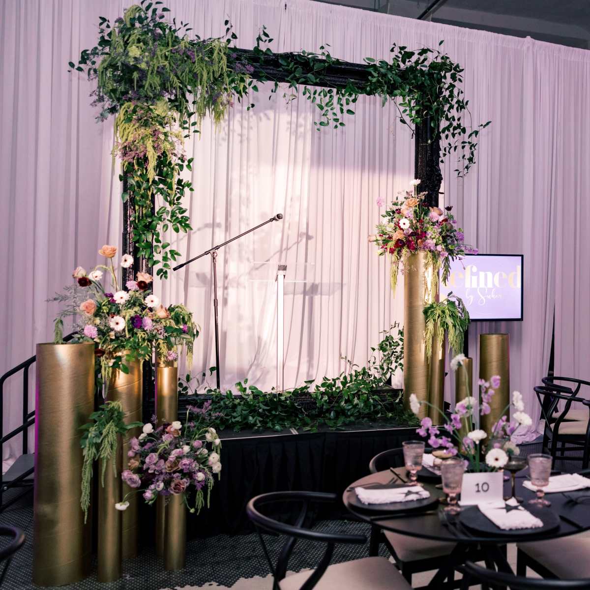 A stage is framed by lush greenery and floral arrangements, with black tables set for dining in the foreground.