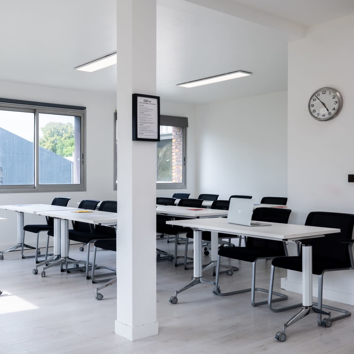 A bright, modern meeting room with a row of tables, black chairs, and large windows.