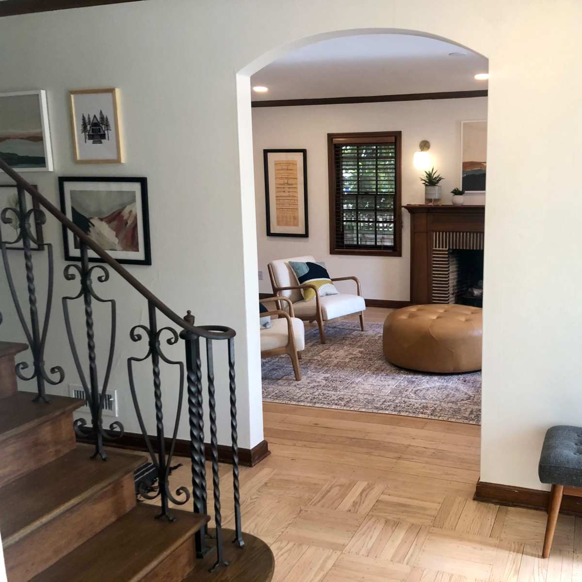 A living space featuring a staircase, a round mirror, and a seating area with two chairs, a large ottoman, and artwork on the walls.