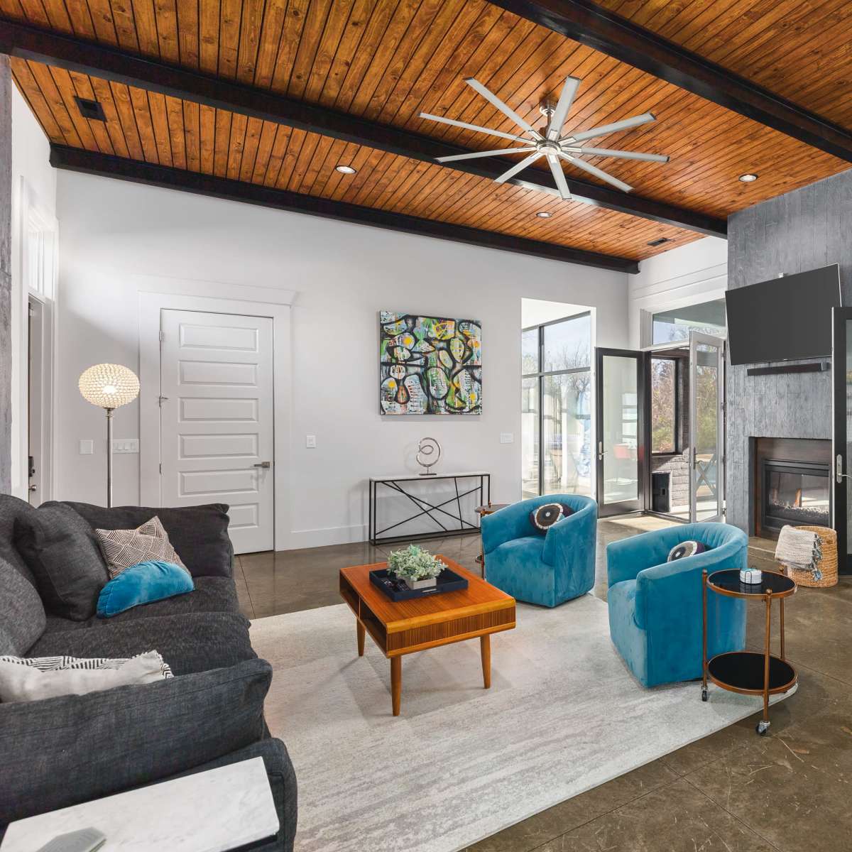 A modern living room with a dark couch, vibrant blue chairs, a wooden ceiling, and large glass doors leading to an outdoor area.