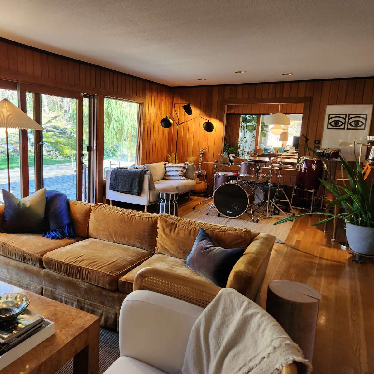 A cozy living room with a brown sofa, a small dining area in the background, and large windows that reveal a view of greenery outside.