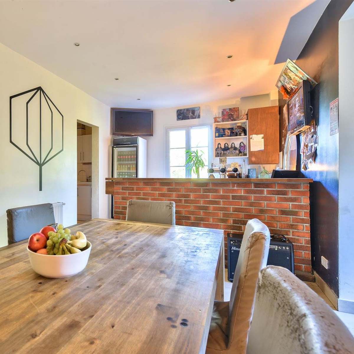 A wooden dining table with a bowl of fruit is set in a kitchen area featuring a brick wall, cabinets, and various decorations.