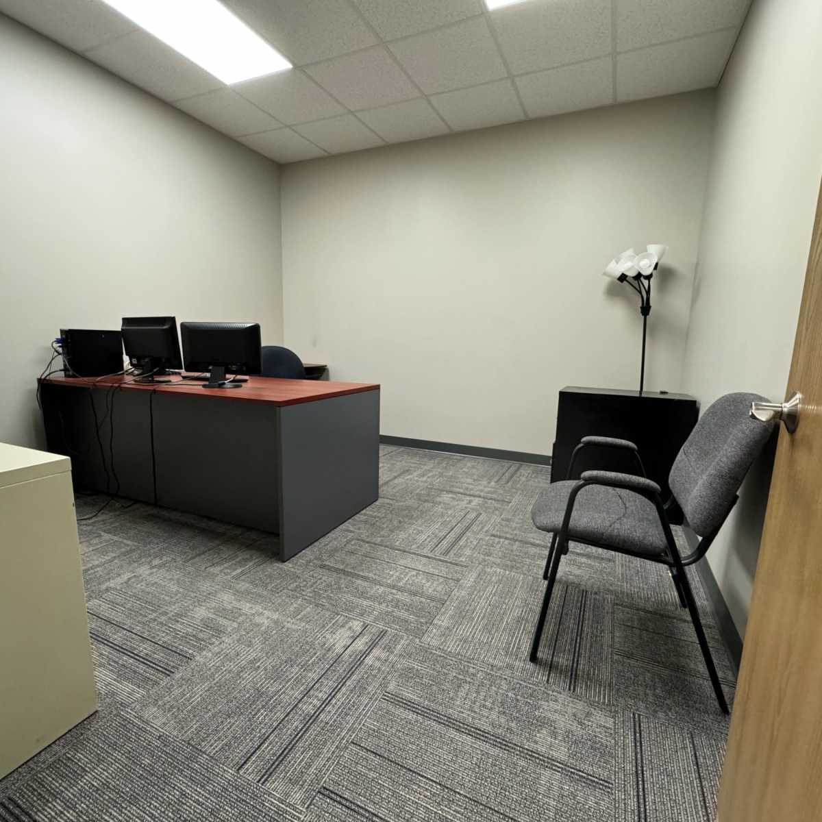 A small, empty office space with a desk, two computer monitors, a chair, and a floor lamp.