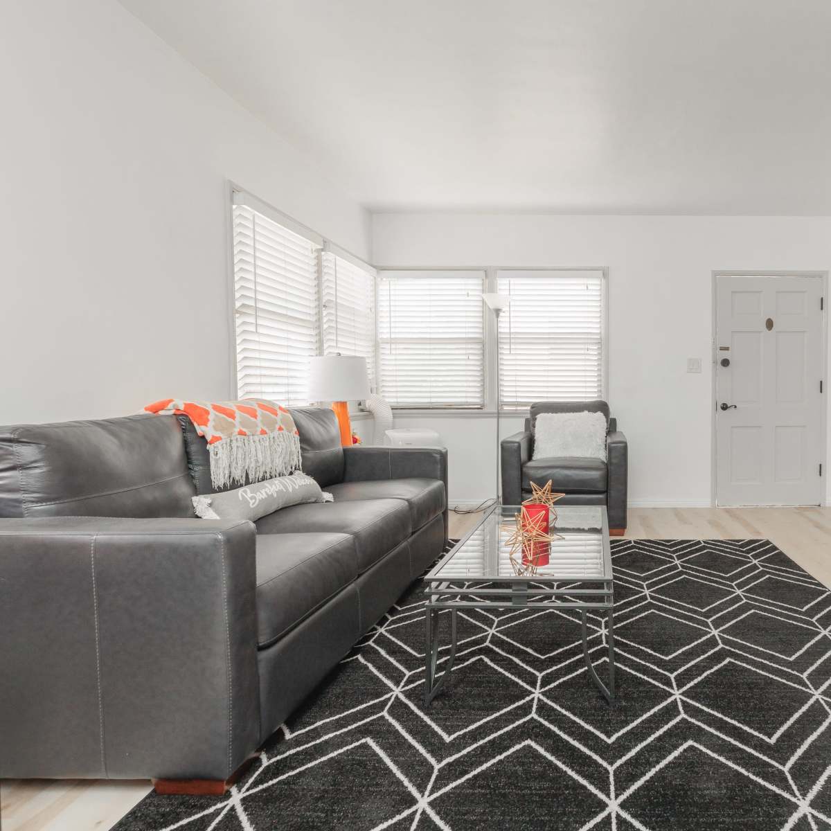 A modern living room with a black leather sofa and armchair, a glass coffee table, and a TV mounted on a wall across from a patterned rug.