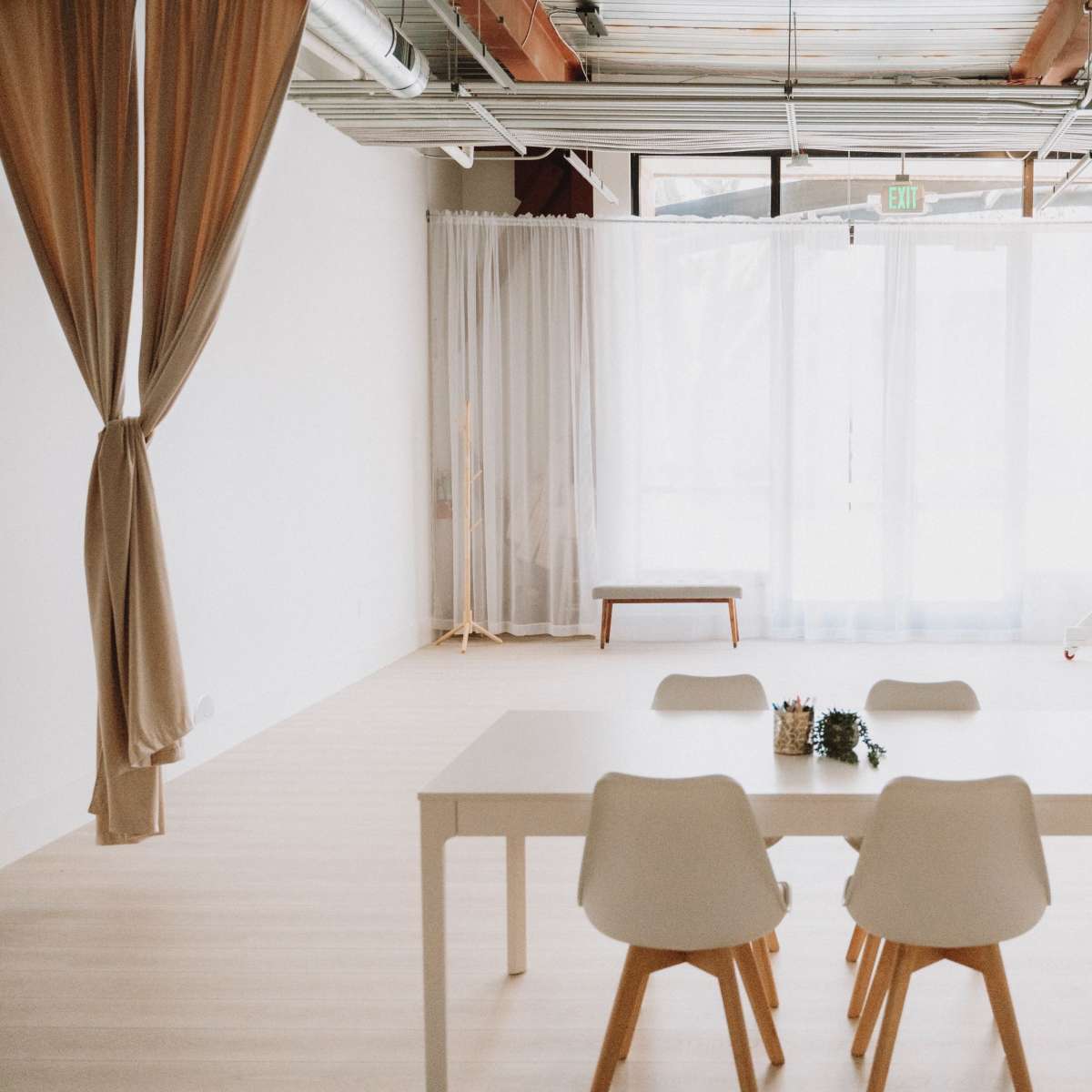 A clean, minimalist room features a white table with four chairs, separated by curtains from a large window and a backdrop on wheels.