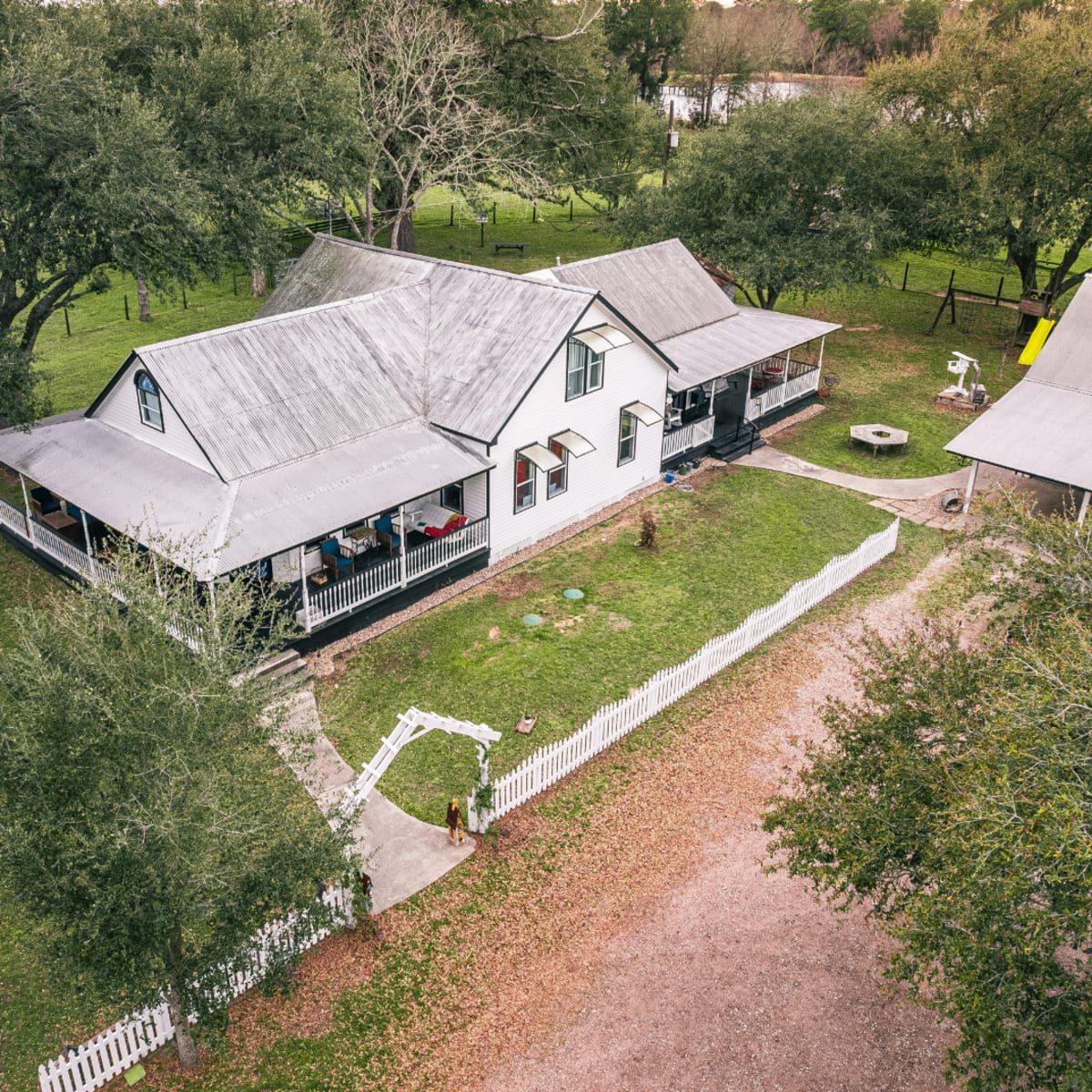 Aerial view of a house with a porch, surrounded by trees and a large grassy yard, with a gravel path and a white picket fence.