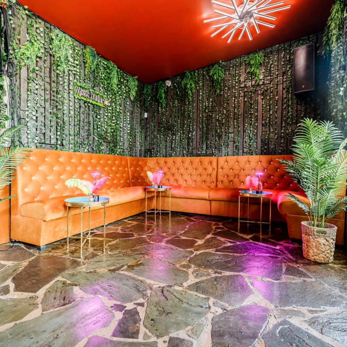 A vibrant lounge area with orange tufted seating, stone flooring, and plants in decorative pots, enhanced by ambient lighting.