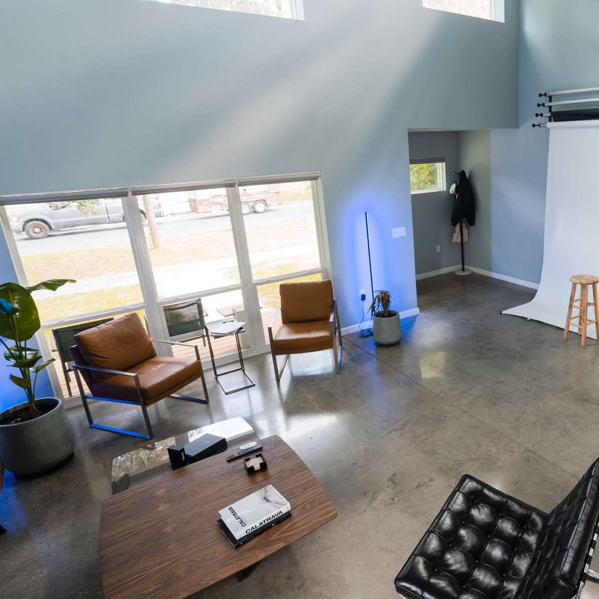 A spacious studio with a white backdrop, a wooden stool, and modern furniture, illuminated by natural light from large windows.