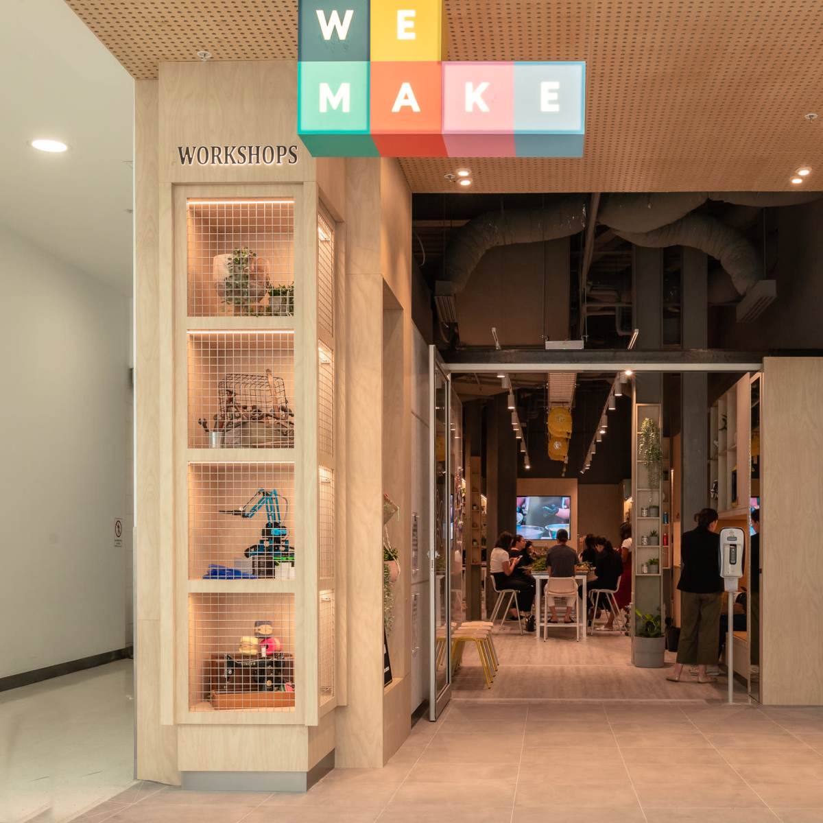A large, modern entrance to a workshop space labeled "WE MAKE," with shelves displaying various items and a seating area visible in the background.