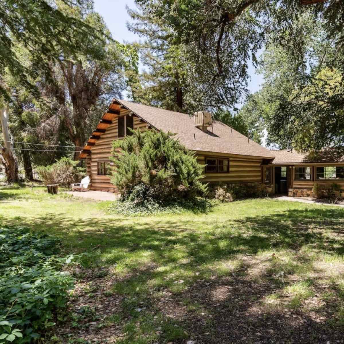 A rustic log cabin with a sloped roof sits among green trees and grassy land.