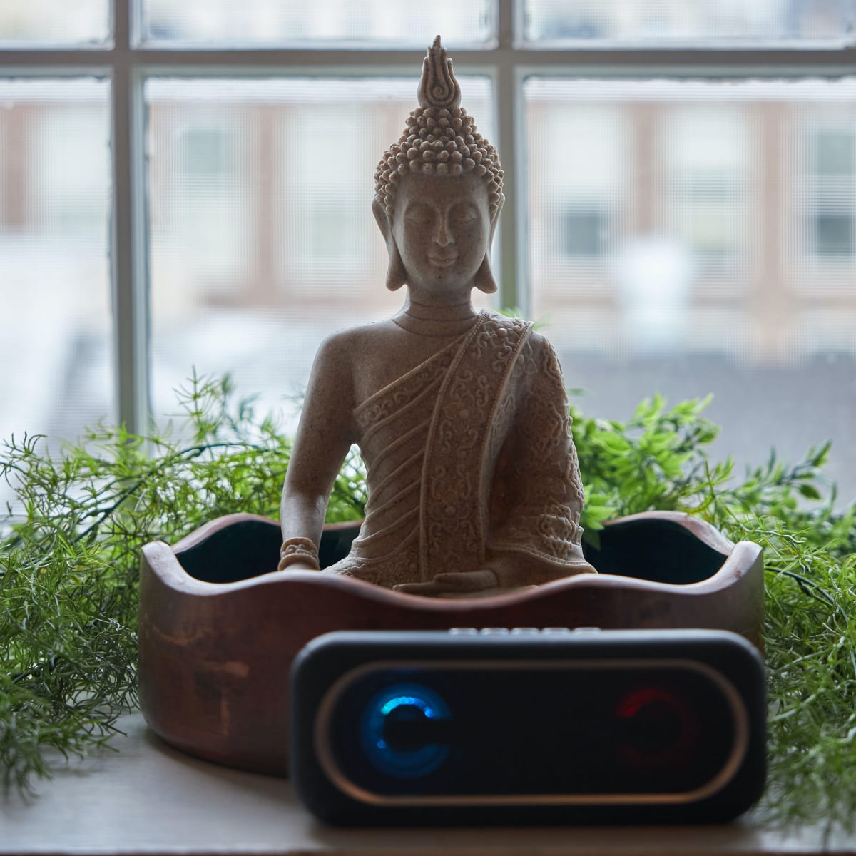 A stone Buddha statue is placed in front of a window, surrounded by greenery and a speaker.