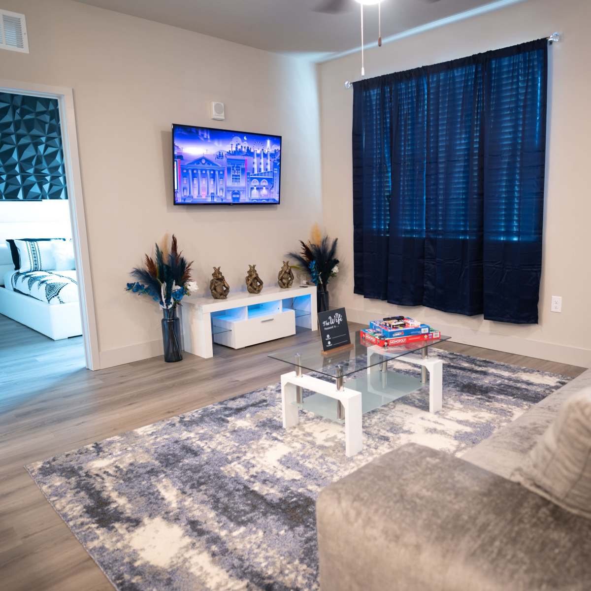 A modern, brightly lit living room featuring a gray sofa, a glass coffee table, and a large television mounted on the wall, with a blue and white color scheme visible in the decor.
