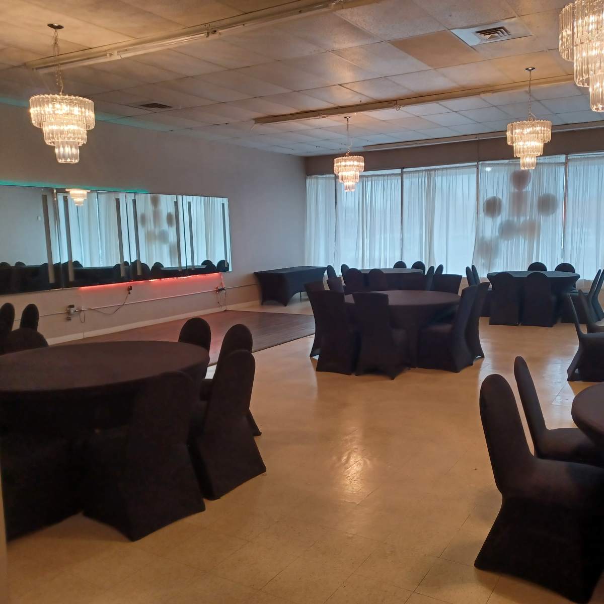 The room features several round black tables surrounded by matching chairs, with chandeliers hanging from the ceiling and a large mirror reflecting the space.