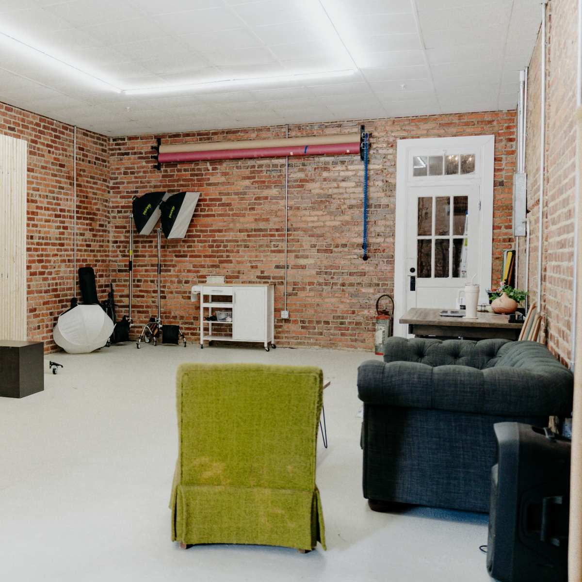 An empty studio space with exposed brick walls, a wooden partition, and furniture including a green chair and a black couch.