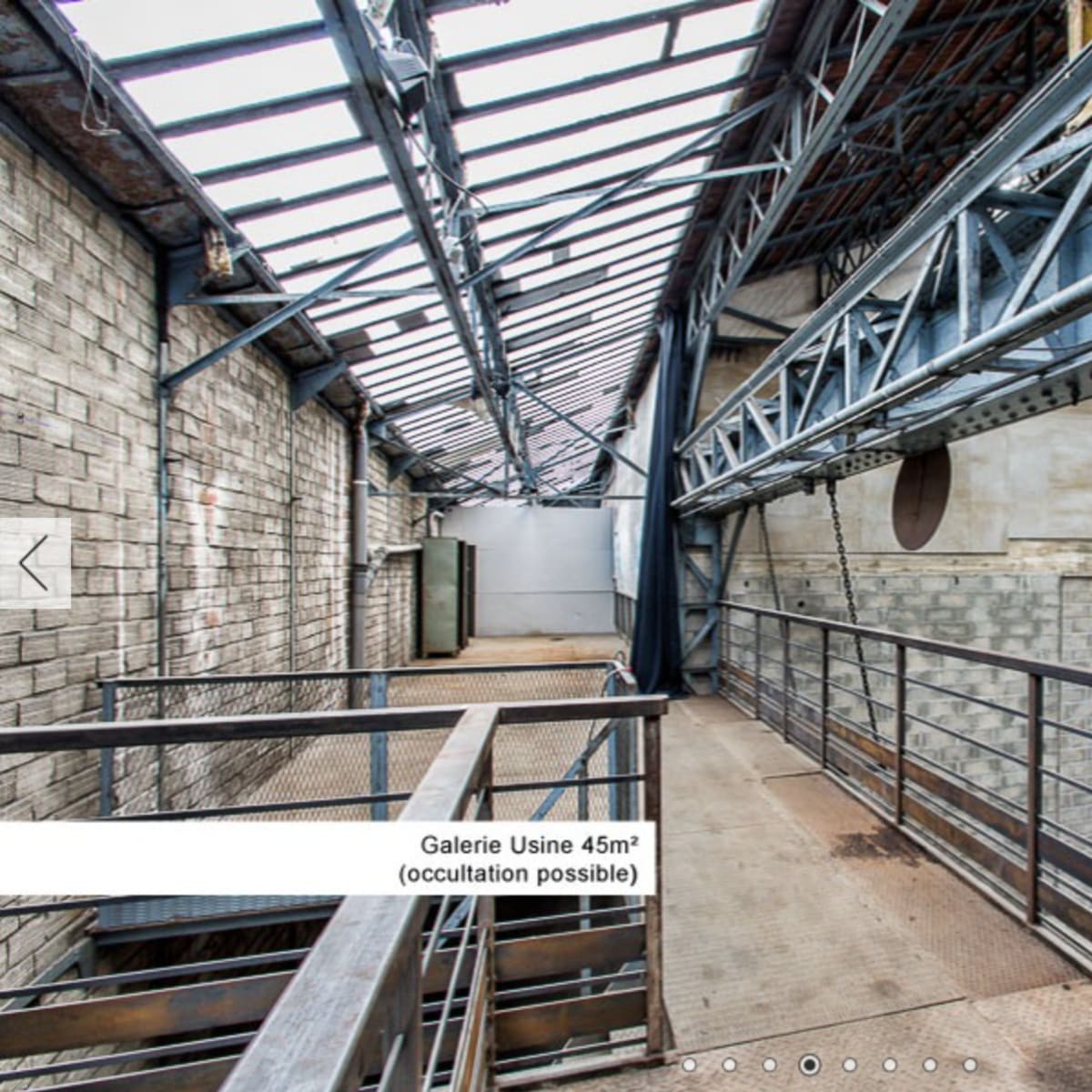 An industrial-style gallery with metal beams and a graffiti-covered wall, featuring a walkway overlooking a spacious area below.