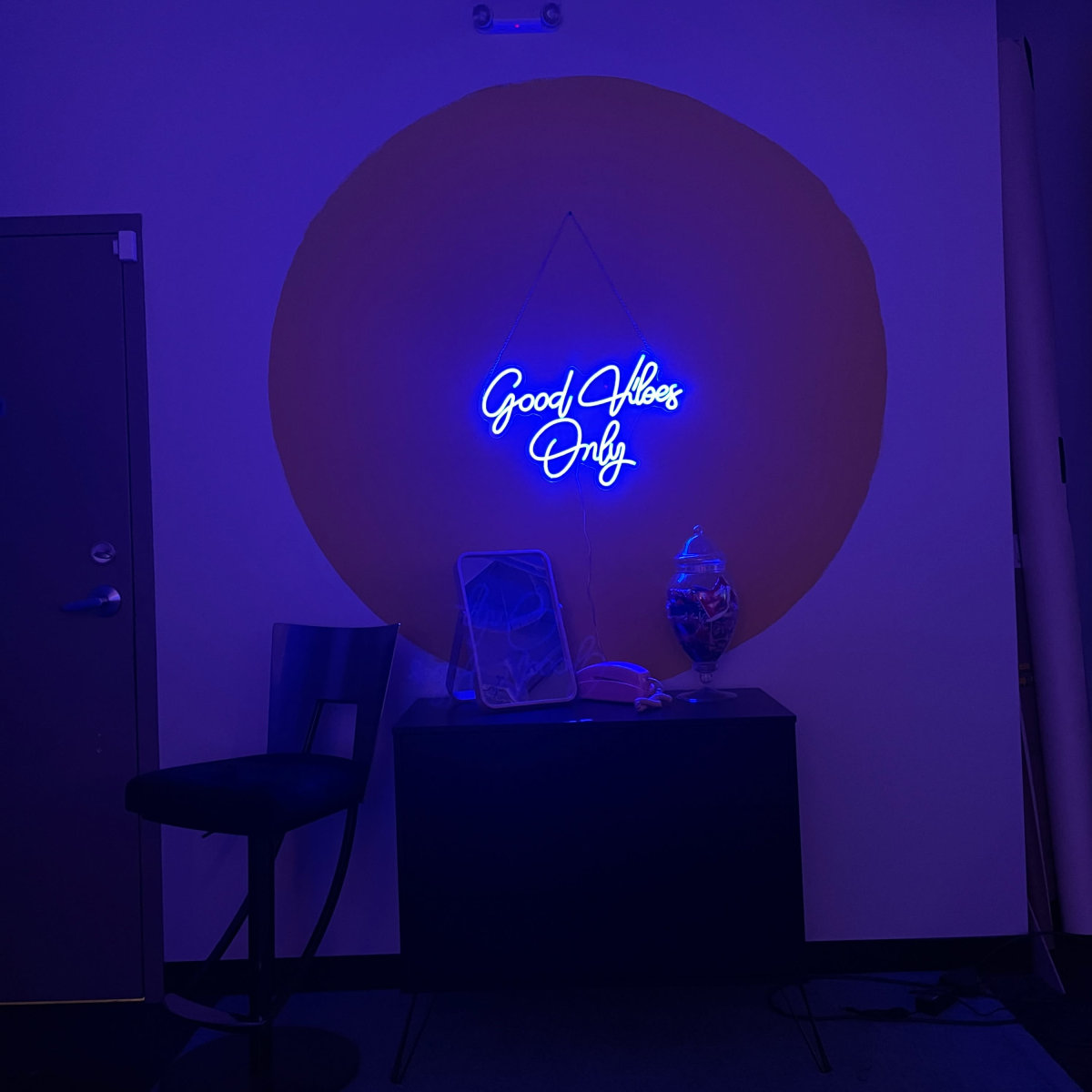A blue neon sign reading "Good Vibes Only" hangs on a yellow circular wall above a small table with a glass container.