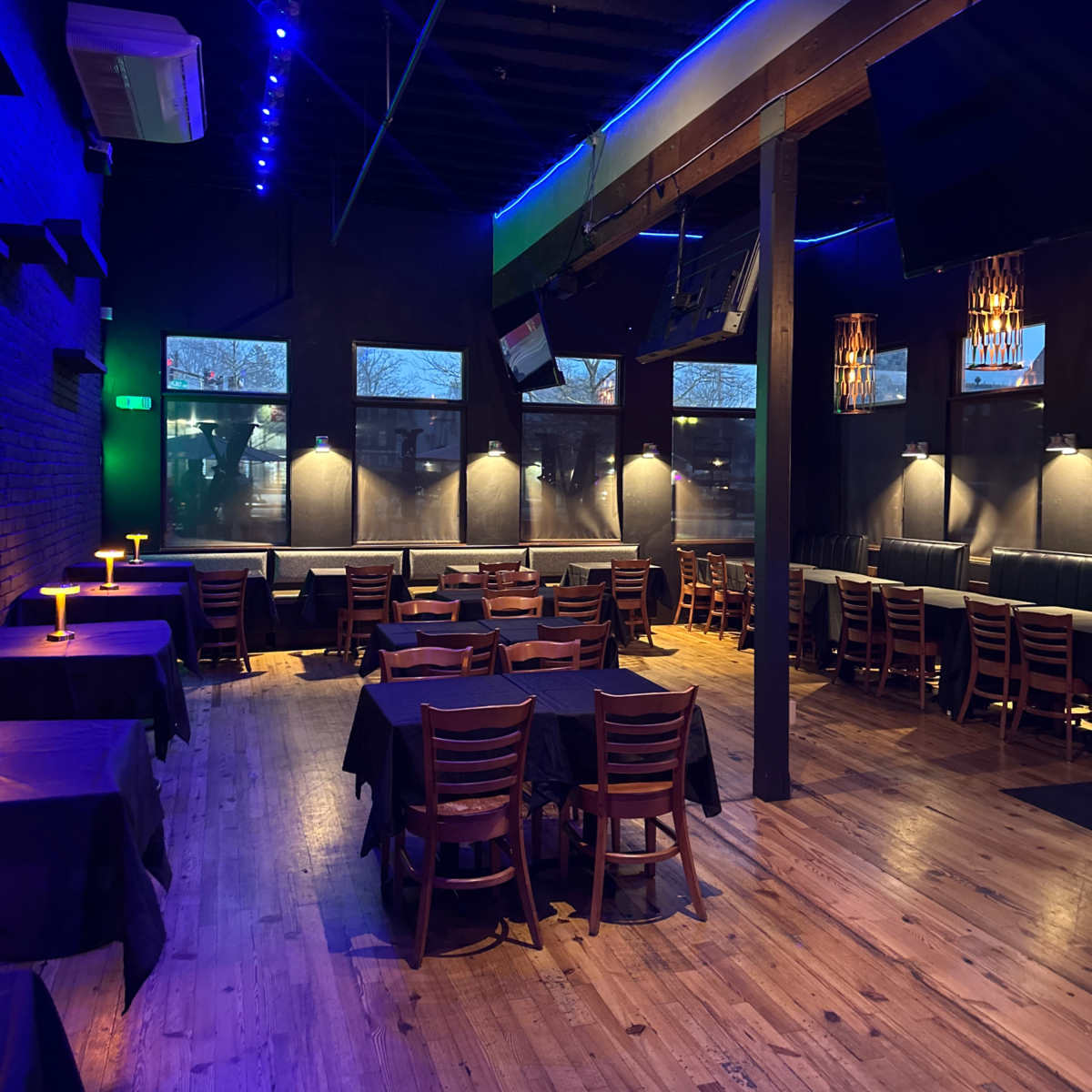 A dimly lit restaurant interior with wooden floors, black tablecloths on tables, and blue accent lighting.