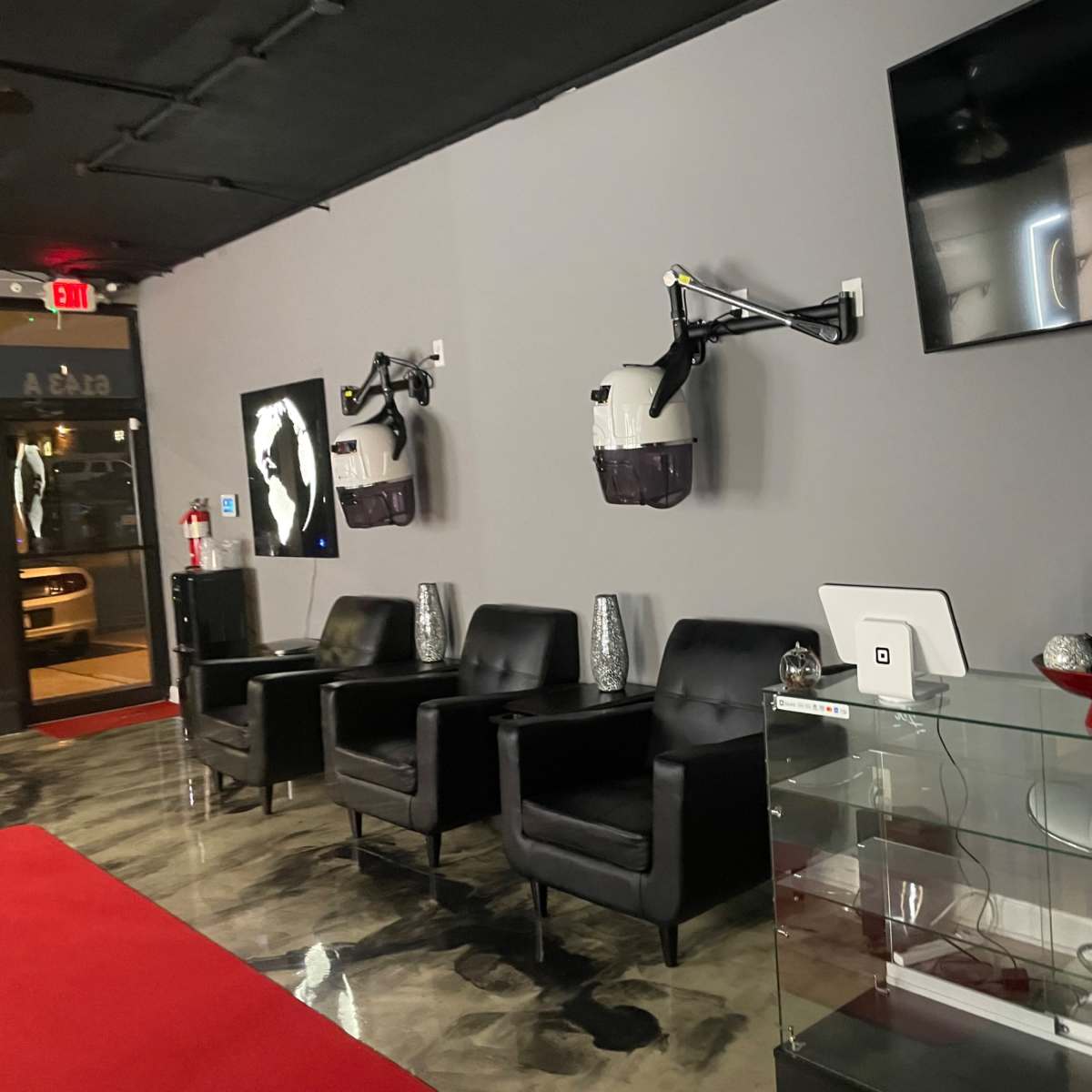 A modern waiting area with black chairs, sleek glass furniture, and a wall-mounted TV, featuring two mounted devices and artwork on the walls.