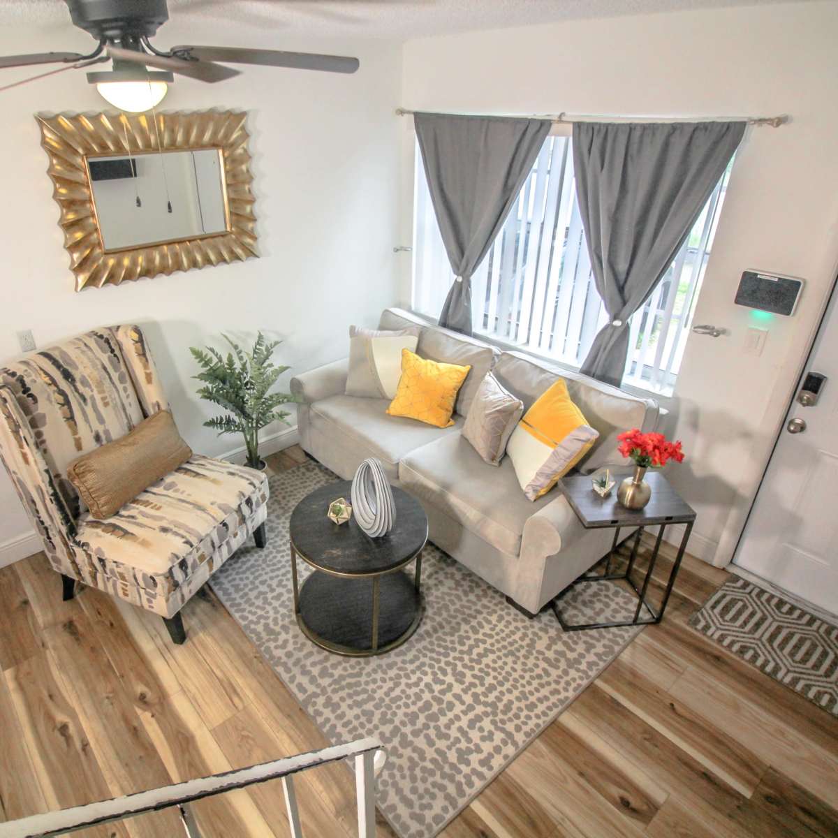A modern living room with a patterned chair, a light-colored sofa adorned with decorative pillows, a coffee table, and a potted plant, all arranged around a window with curtains.