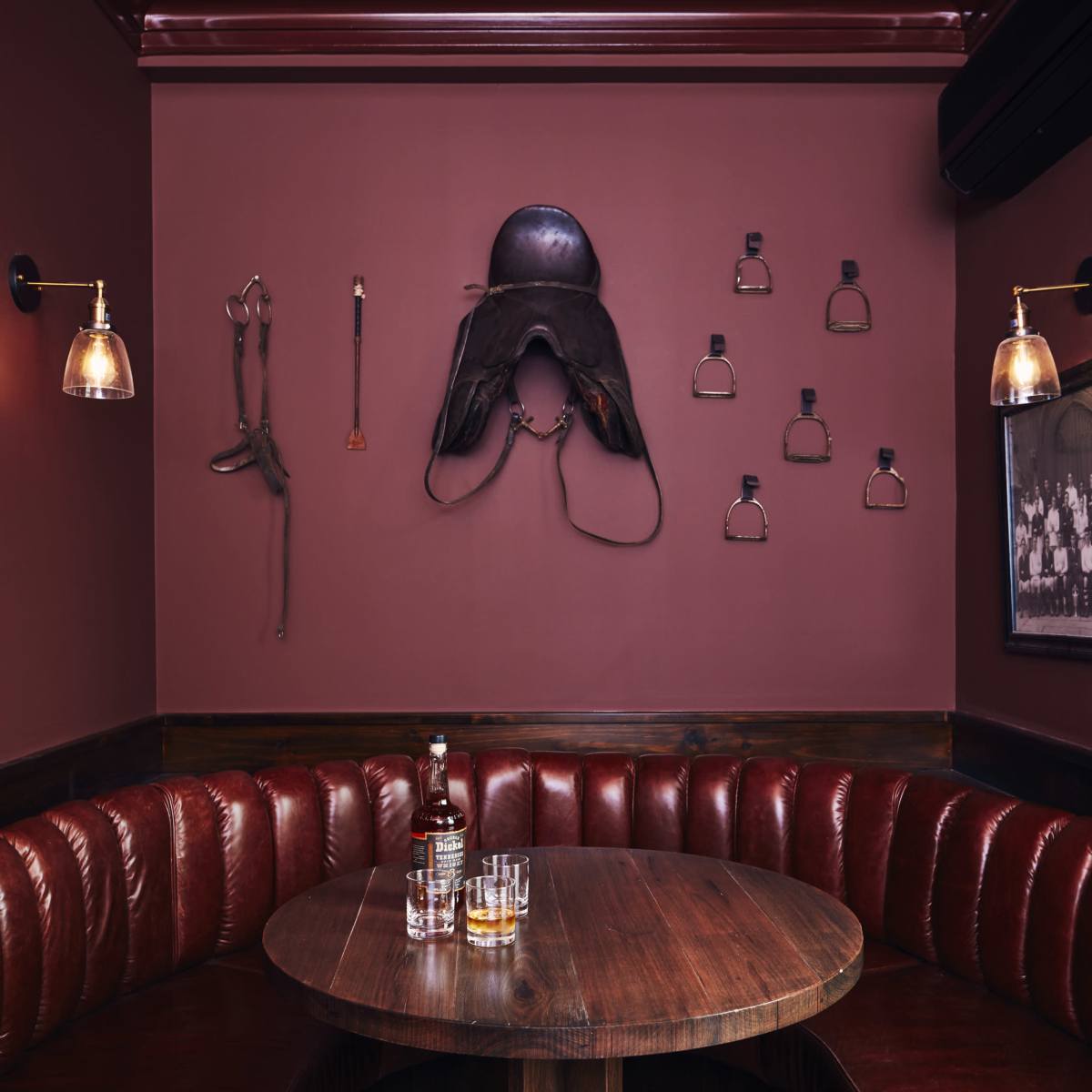 A cozy booth in a dimly lit restaurant features a round wooden table, surrounded by padded red leather seating, with equestrian-themed decor on the wall.