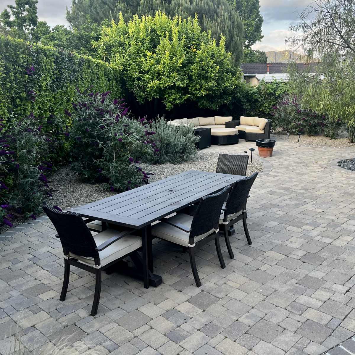 A paved patio area features a rectangular table with chairs, surrounded by garden plants and a sectional seating arrangement.