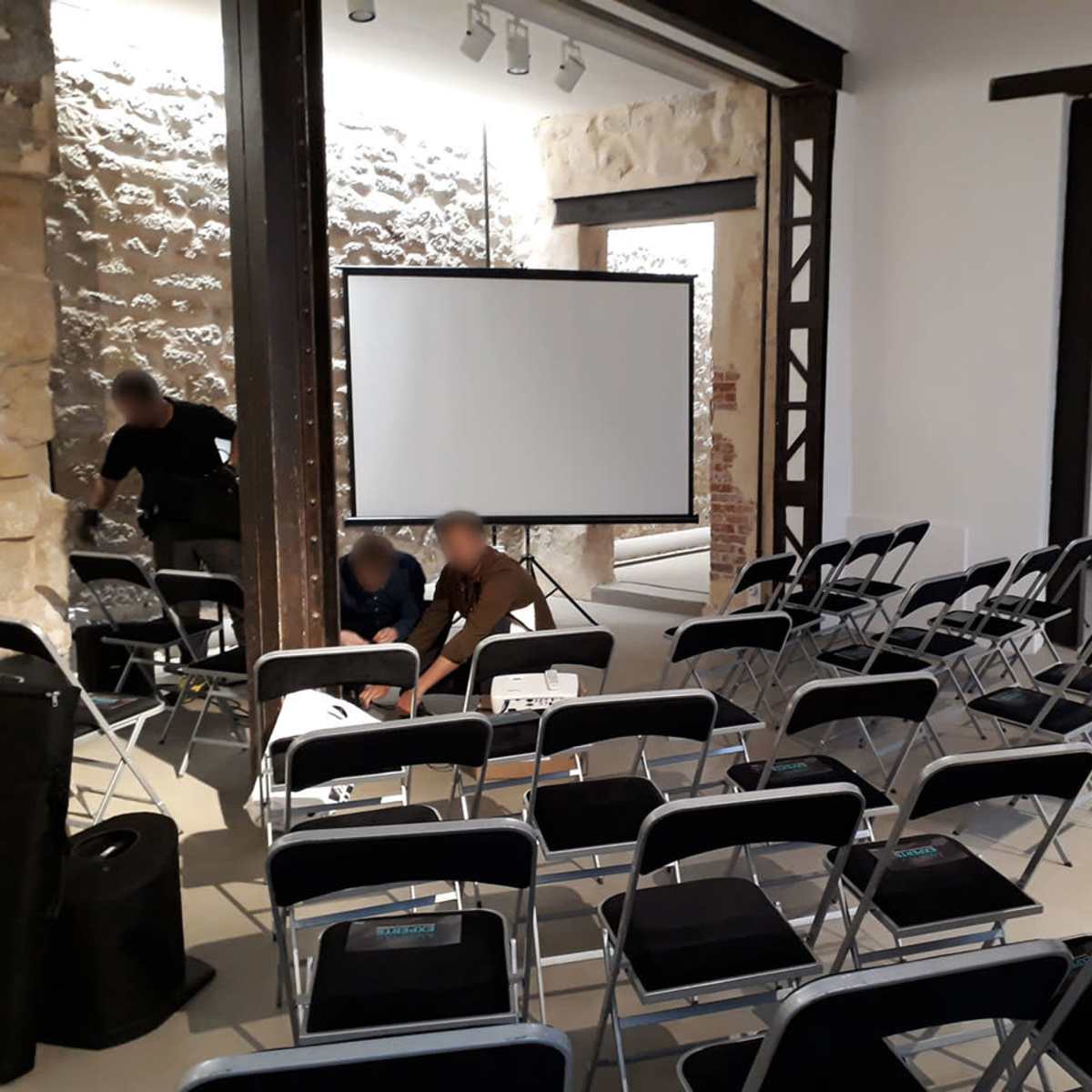 A small group is preparing a meeting space with rows of chairs facing a projector screen in a room with exposed stone walls and wooden beams.