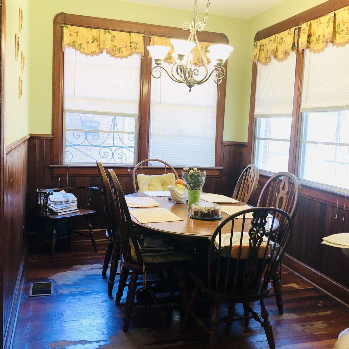 A dining room features a wooden table surrounded by chairs under a chandelier, with windows adorned with floral valances.