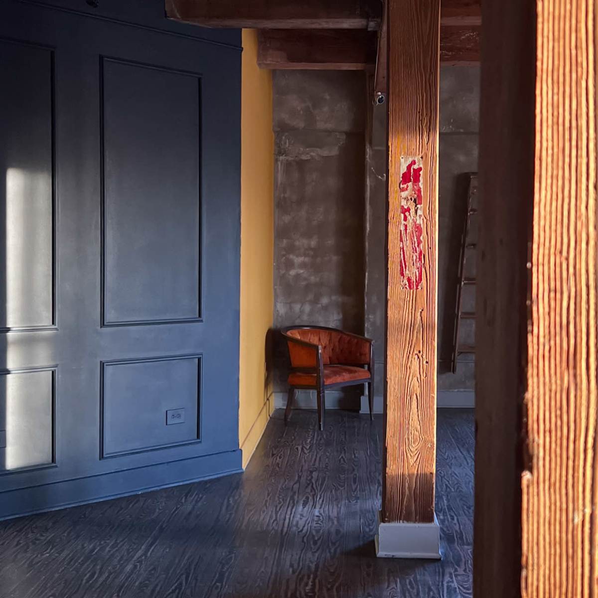 A partially furnished room with wooden beams, a yellow wall, and a brown chair in the corner.