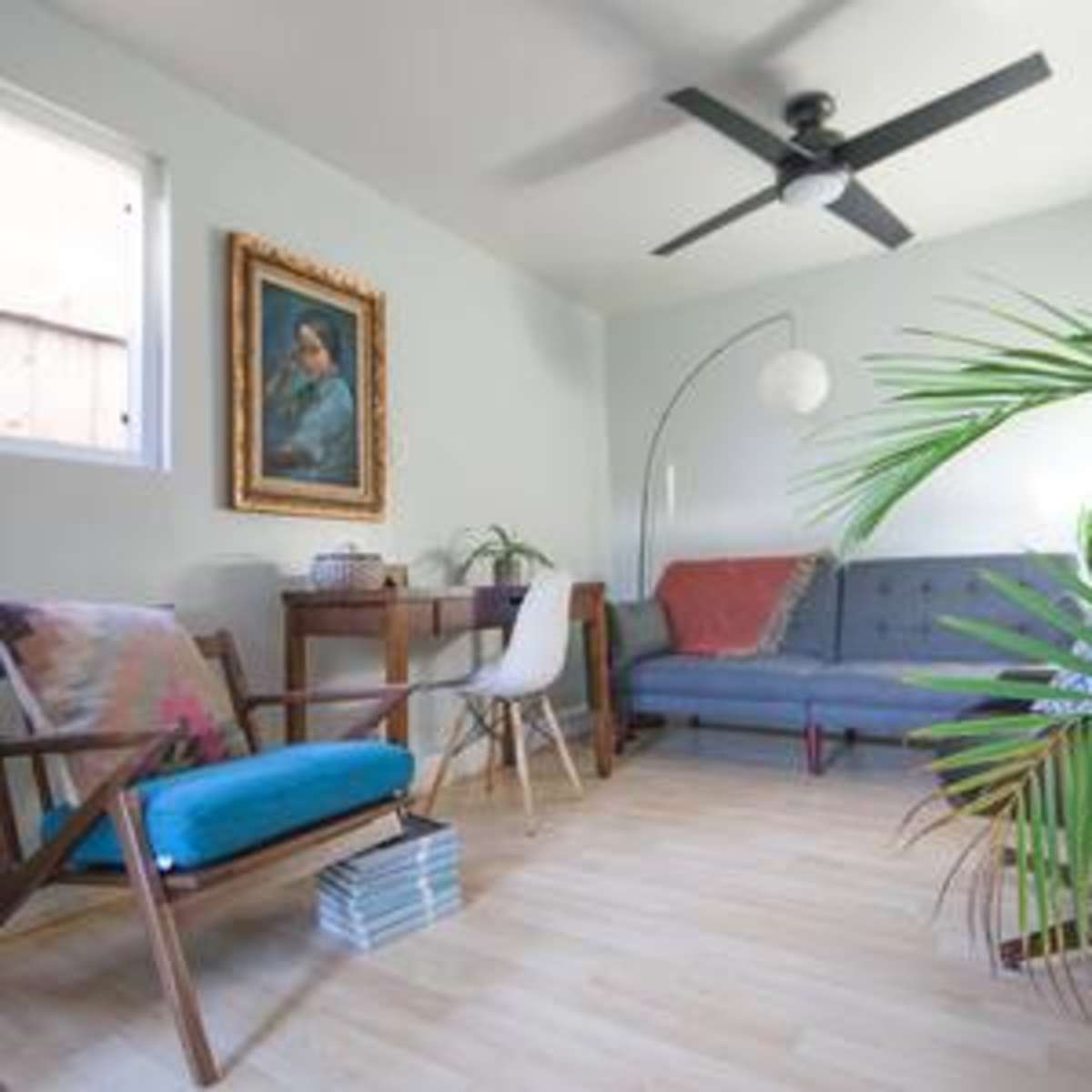 A well-lit living room featuring a gray sofa, a wooden chair with a blue cushion, a floor lamp, and several potted plants.