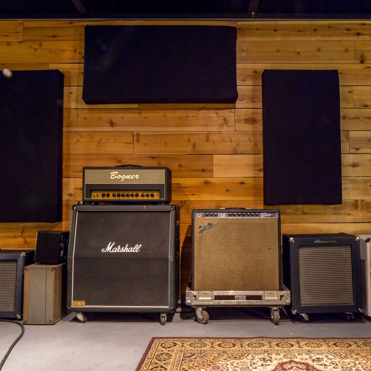 A wall of music amplifiers arranged on a wooden floor with acoustic panels mounted on the wall.