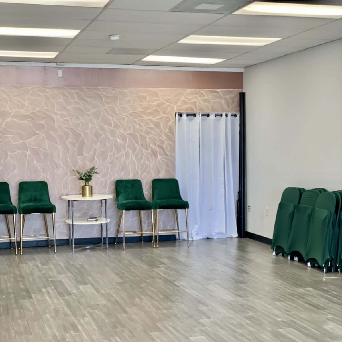 A spacious room with a light-colored textured wall, three green chairs with gold legs, a small round table with a plant, and a stack of green chairs against the wall.