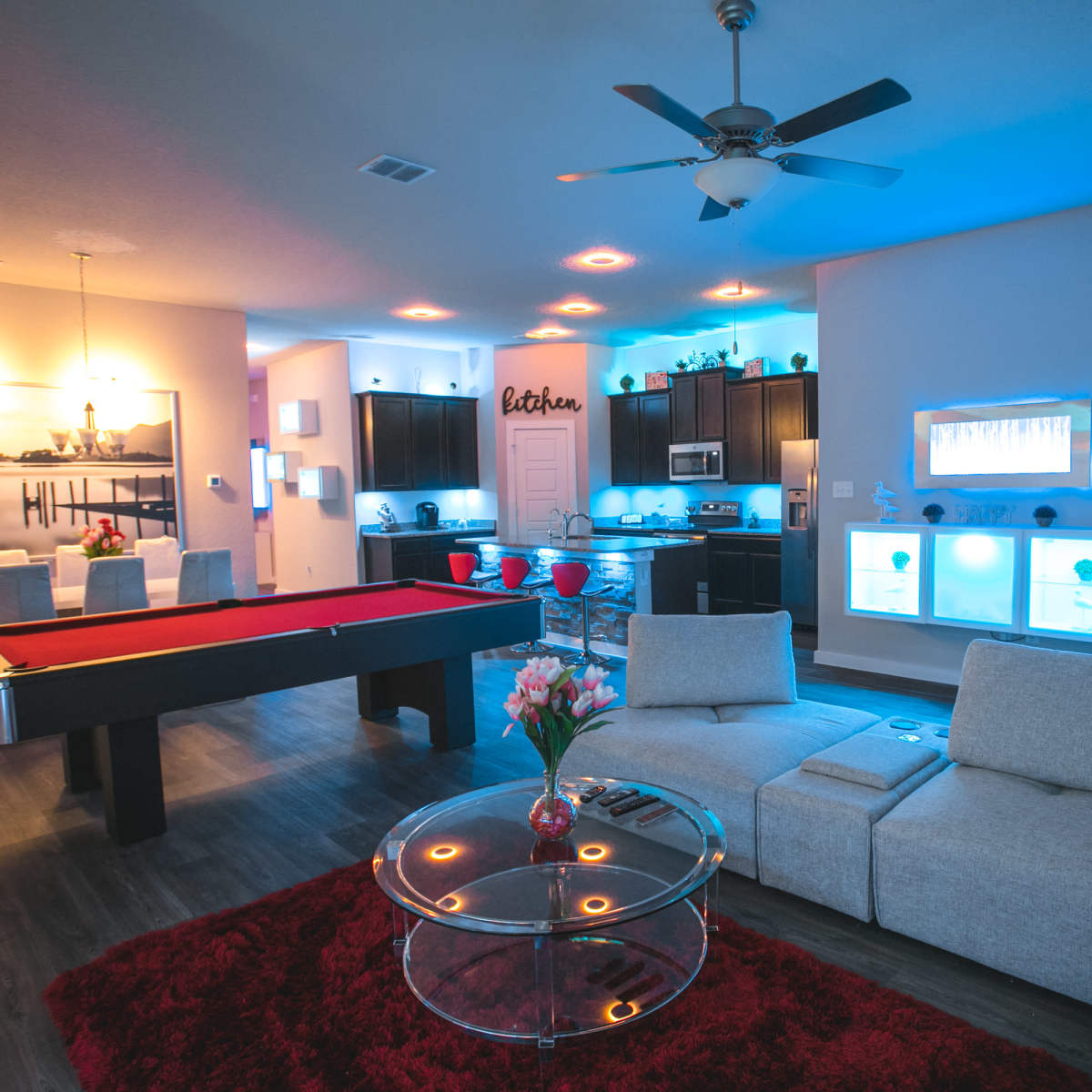 A modern living room with a pool table, a sectional sofa, and a kitchen area illuminated with blue lighting.