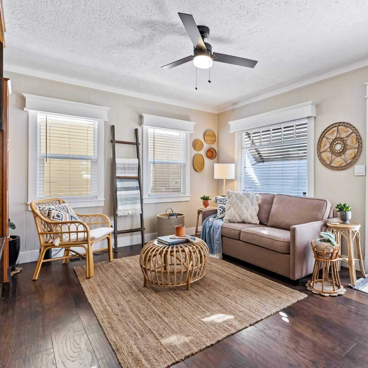 The room features a light-colored living area with a sofa, a round woven coffee table, and large windows allowing natural light to enter.