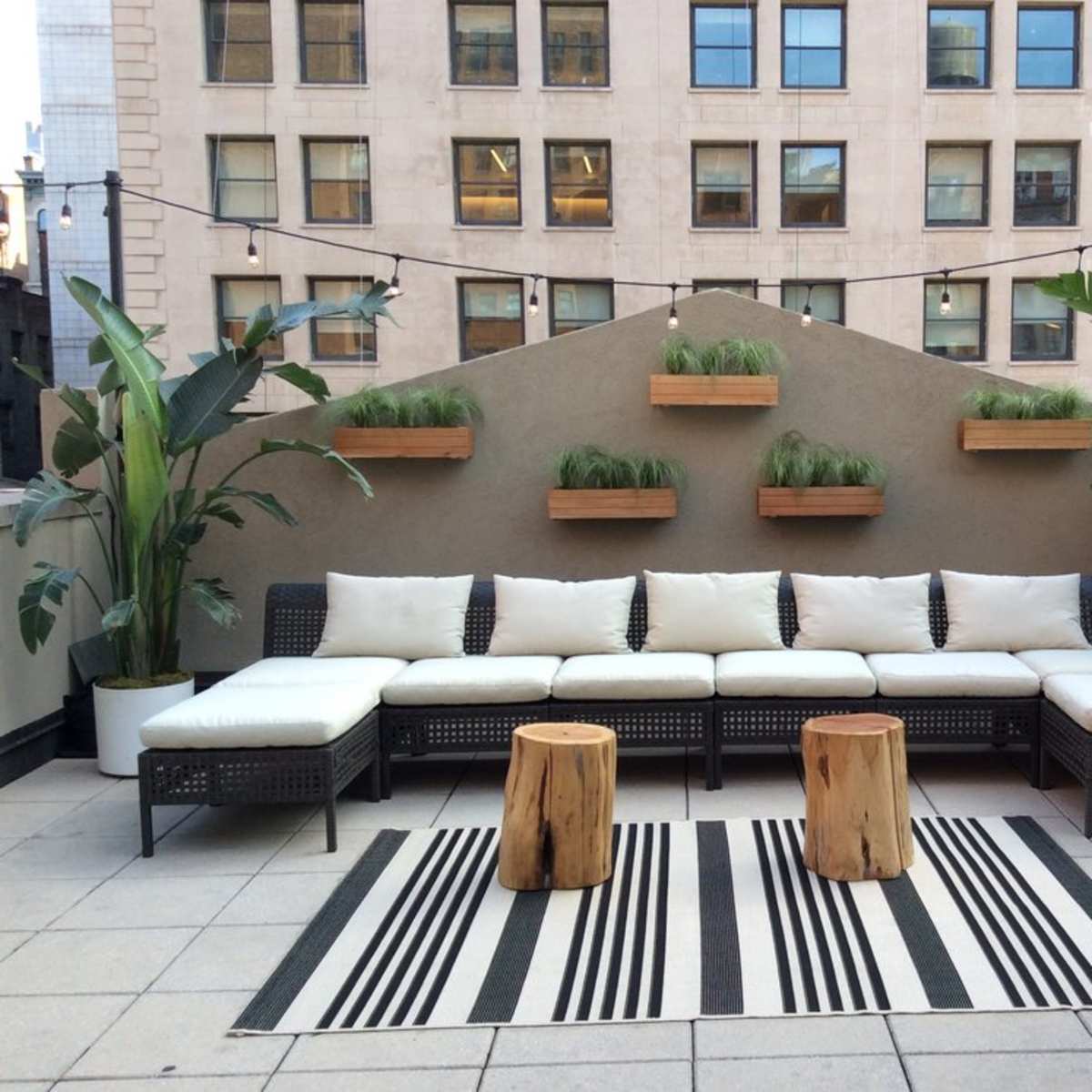 A rooftop terrace with a sectional sofa adorned with white cushions, two wooden stools, and wall-mounted planters filled with greenery.