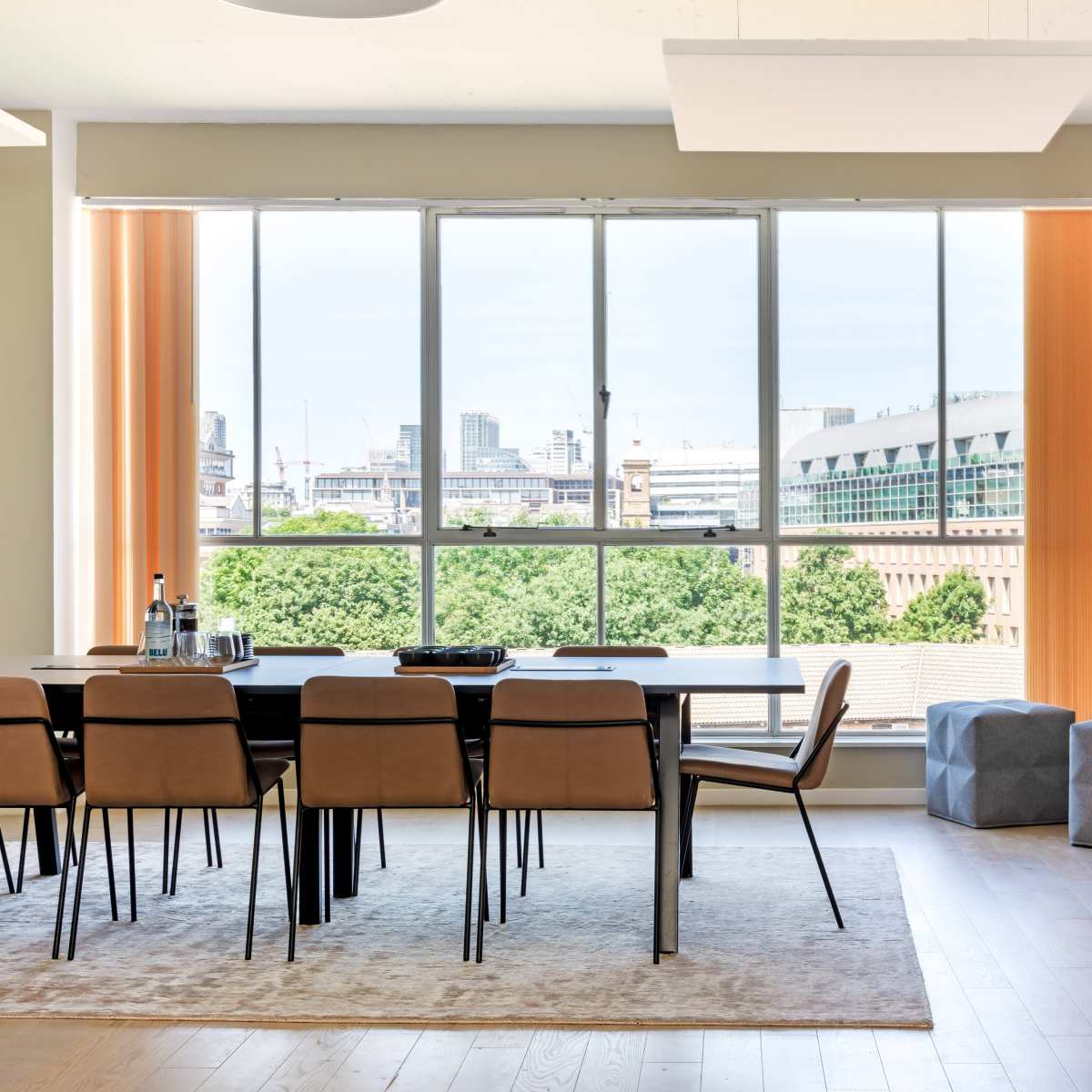 A modern dining area features a long table surrounded by chairs, with large windows offering a view of the city skyline.
