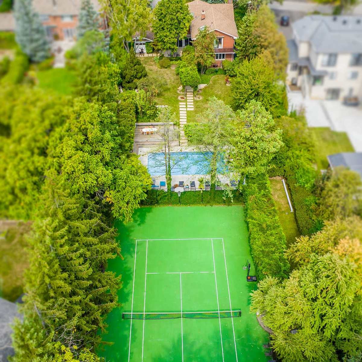 An aerial view of a home with a green tennis court surrounded by trees, a swimming pool, and a pathway leading to the house.
