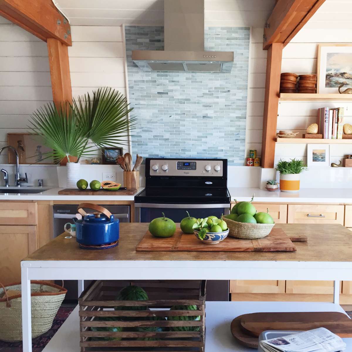 A modern kitchen features a central island with green apples, a stove, wooden shelves, and decorative plants in a bright, airy space.